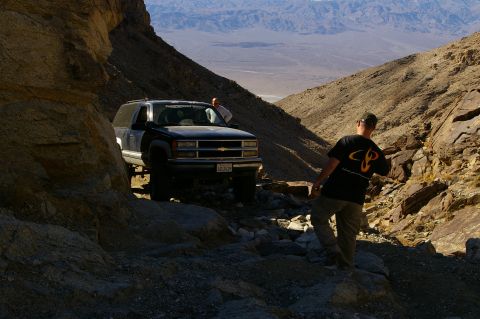1986 Ford Bronco - Chevrolet Suburban driving on a rough off-road trail in a desert canyon.
