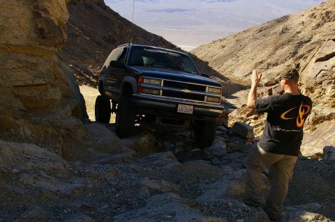 1986 Ford Bronco - A person directs a lifted Chevrolet Tahoe on a challenging off-road path in a desert canyon.