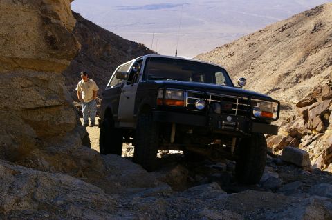 1986 Ford Bronco - A two-tone Ford Bronco navigates a , rocky desert path, with a man observing from the side.