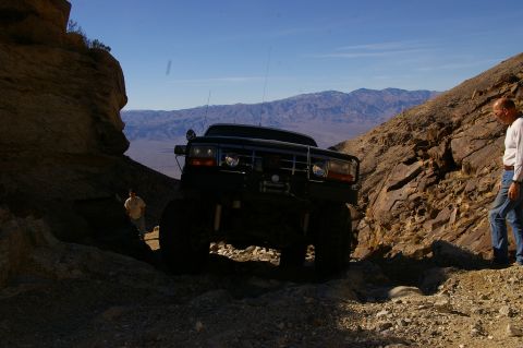 1986 Ford Bronco - A man stands by a black Ford F-Series truck navigating a narrow, rocky canyon with mountains in the distance.