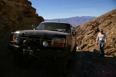 1986 Ford Bronco - A man walks beside a black Ford F-Series truck navigating a steep, rocky trail with mountains in the background.