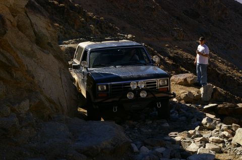 1986 Ford Bronco - A 1980s Ford Bronco navigates a challenging, rocky off-road path with a person observing from the side.