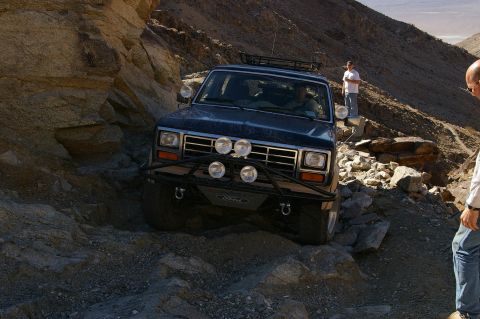 1986 Ford Bronco - A dark blue 1986 Ford Bronco navigates a challenging rocky terrain, equipped with an aftermarket bumper and auxiliary lights.