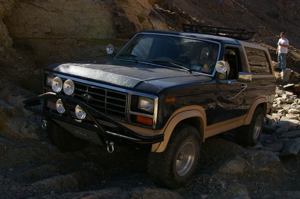 Dark blue and tan Ford Bronco with aftermarket bumper and lights, driving on rocky terrain