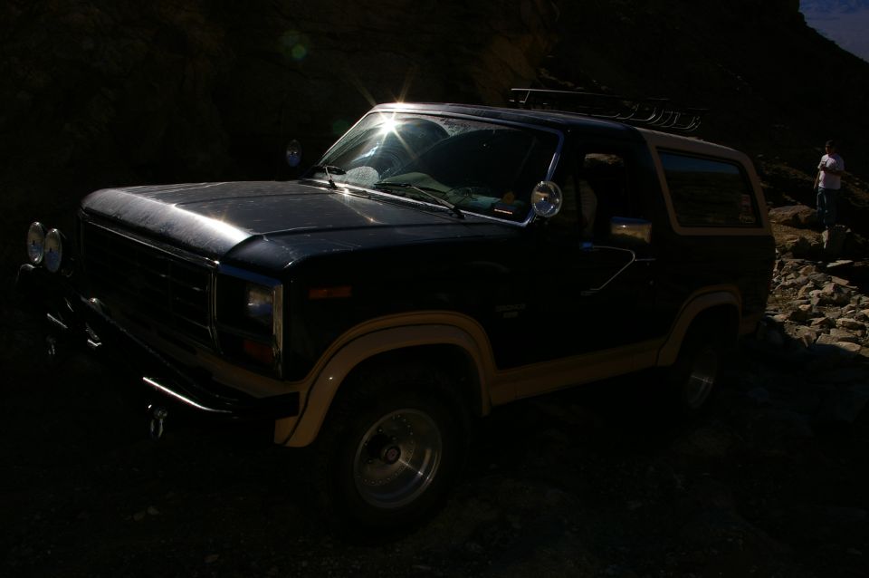 1986 Ford Bronco with tan trim parked on a rocky, sunlit path. A person stands on rocks in the background