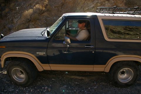 1986 Ford Bronco - A man drives a classic Ford Bronco through a rocky, arid landscape, reflecting the surrounding hills in its windows.