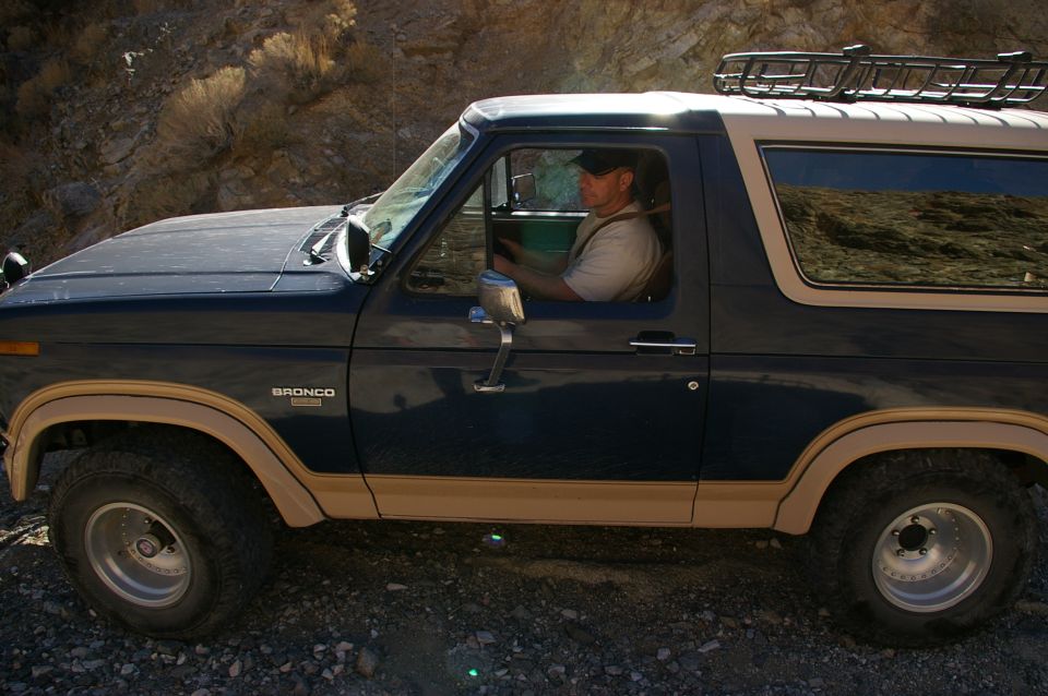 Side view of a dark blue Ford Bronco with tan trim, driven by a man in a cap. It's on a dirt road with rocky hills in the background