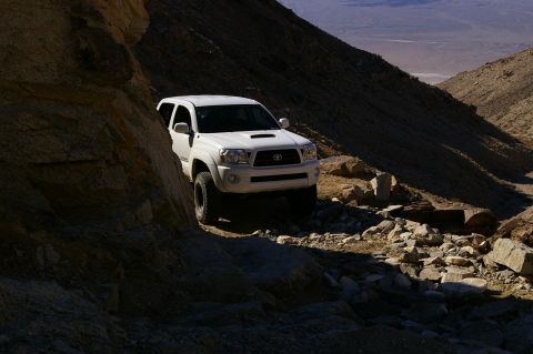 1986 Ford Bronco - White Toyota Tacoma TRD Off-Road pickup truck navigates a , rocky desert trail under bright sunlight.