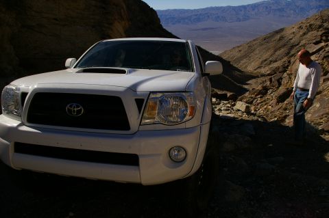 1986 Ford Bronco - A man stands next to a white Toyota Tacoma on a desert path with mountains in the background.