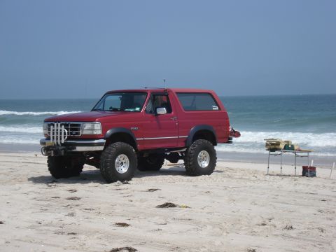 1994 Ford Bronco - Red lifted full-size Ford Bronco with hardtop sitting on the beach with breaking surf behind it, shown from the driver's side slightly forw…