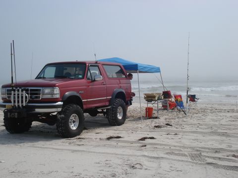 1994 Ford Bronco - Red lifted full-size Ford Bronco with hardtop parked on foggy beach sand, viewed from the driver's-side 3/4 front with mud-terrain tires on…