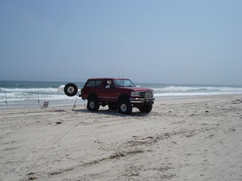1994 Ford Bronco - Red lifted Ford Bronco parked on a wide sand beach at the surf line under a hazy blue sky, seen from the front three-quarter passenger side…