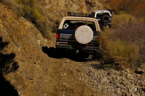 1986 Ford Bronco - Two SUVs navigate a steep, rocky mountain path with sparse vegetation.