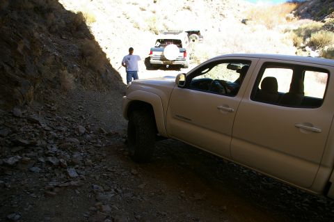 1986 Ford Bronco - Two vehicles navigate a tight, sun-dappled canyon trail with a lone figure observing their progress.