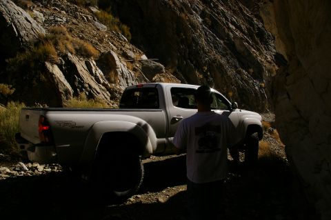 1986 Ford Bronco - A white Toyota Tacoma TRD Sport navigates a tight, rocky mountain pass, with a person standing nearby.