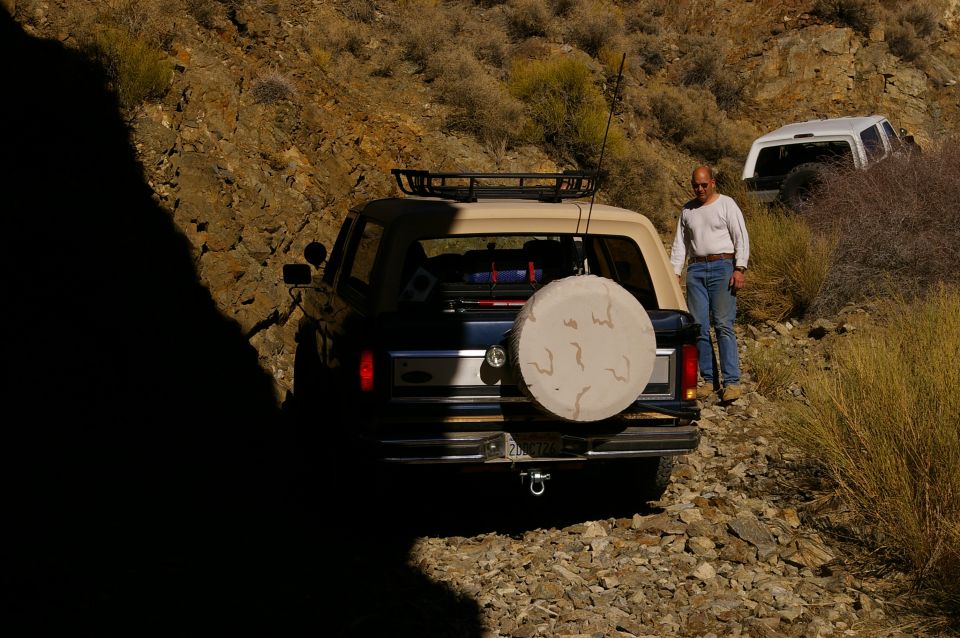 1986 Ford Bronco parked on a rocky trail with a man standing beside it; another white SUV is visible further up the trail