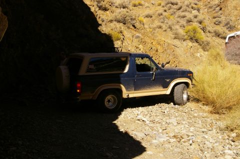 1986 Ford Bronco - A Ford Bronco navigates a narrow, rocky path with dry brush and a second vehicle visible in the distance.
