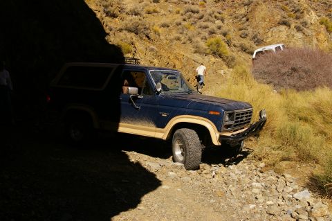 1986 Ford Bronco - A dark blue Ford Bronco navigates a rough, rocky terrain with another white vehicle visible in the background.