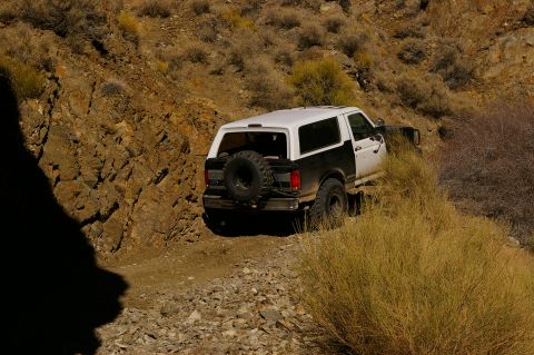 1986 Ford Bronco - A two-tone Ford Bronco navigates a rocky, off-road trail with sparse vegetation.