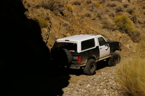 1986 Ford Bronco - A two-tone Ford Bronco navigates a rough, rocky terrain under a bright sky.