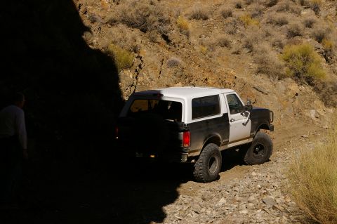 1986 Ford Bronco - A two-tone Ford Bronco navigates a , off-road trail under a bright sky.