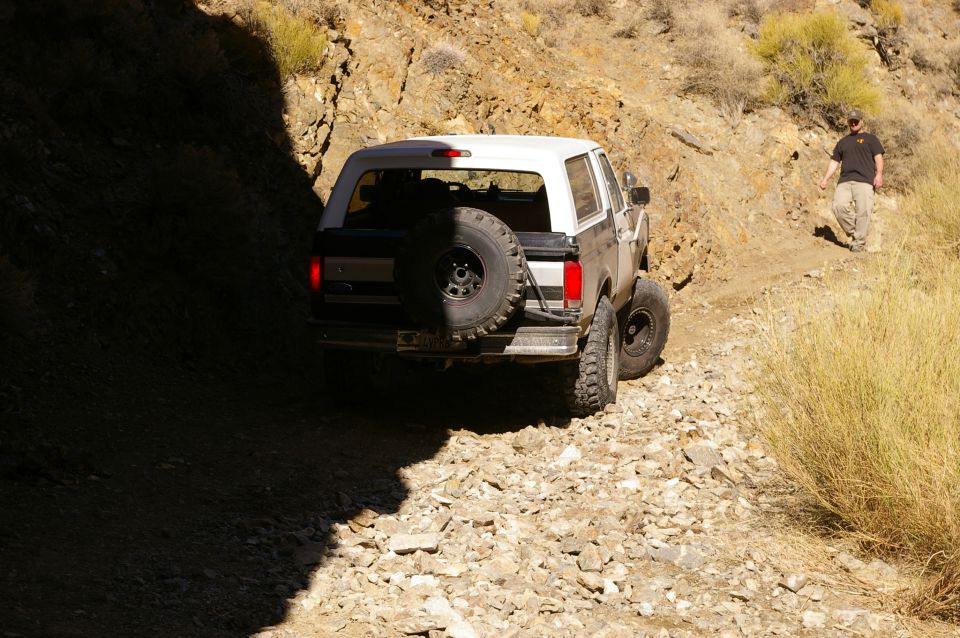 1986 Ford Bronco driving on a rough, rocky path with a person walking behind