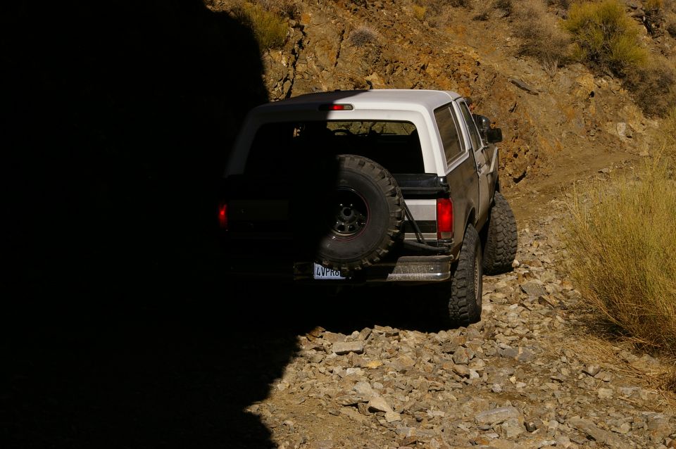 Ford Bronco on a rocky, off-road trail, partially in shadow. Spare tire mounted on the back