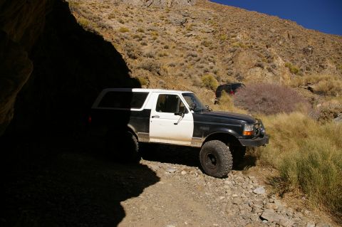 1986 Ford Bronco - A two-tone Ford Bronco is parked on a rough trail, partially in shadow, with another vehicle in the distance.