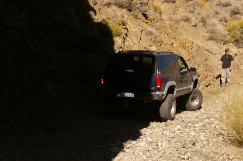 1986 Ford Bronco - Black Chevrolet Tahoe navigating a rough, rocky trail in a desert landscape.