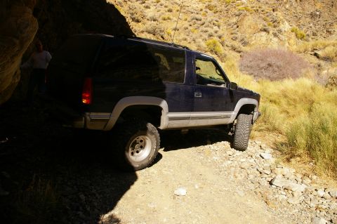 1986 Ford Bronco - A dark blue SUV navigates a rough, rocky terrain, partially shaded by a large rock formation.