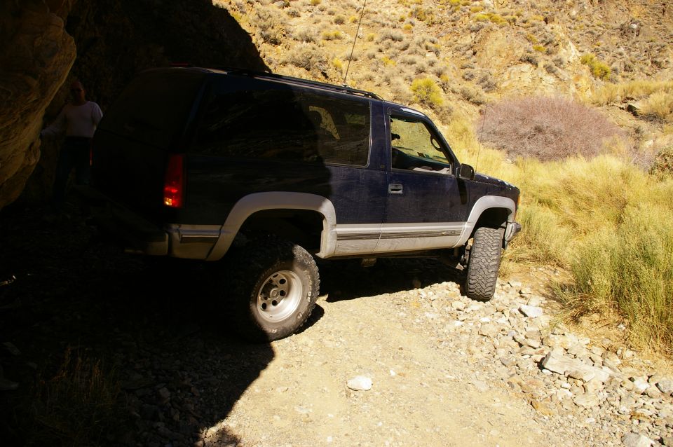 Dark blue SUV parked on a rocky, off-road trail next to a large rock overhang. A person stands near the driver's side rear