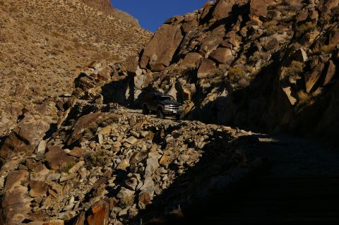1986 Ford Bronco - An SUV drives along a treacherous, rocky path in a mountainous desert landscape.