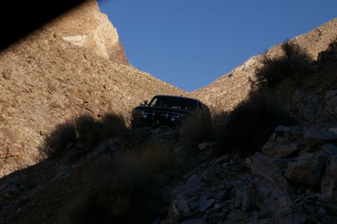 1986 Ford Bronco - A dark SUV navigates a narrow, rocky canyon under a clear blue sky.