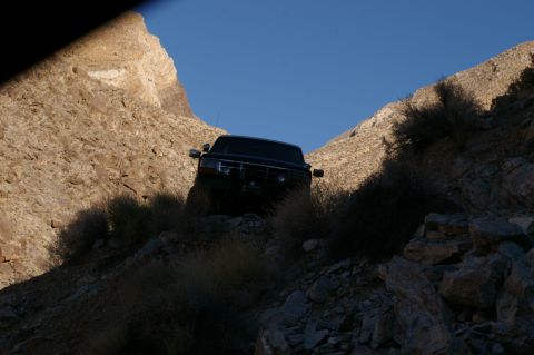 1986 Ford Bronco - A dark SUV navigates a challenging, narrow off-road path between rocky hills under a clear blue sky.