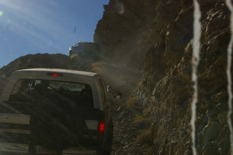 1986 Ford Bronco - A white 1986 Ford Bronco navigates a narrow, dusty mountain road, with another vehicle visible ahead on the trail.