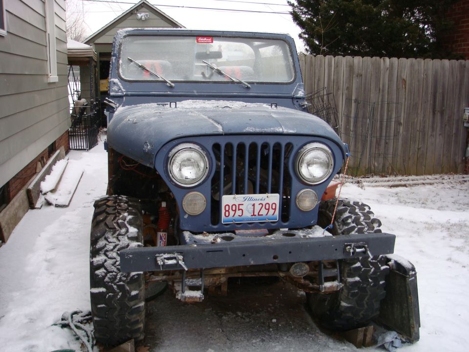 Front view of a blue Jeep CJ parked in the snow. It has large tires and a license plate reading '895 1299'