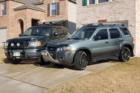 2006 Ford Escape - Two SUVs, a 2006 Ford Escape and a black SUV, are parked in front of a brick house.