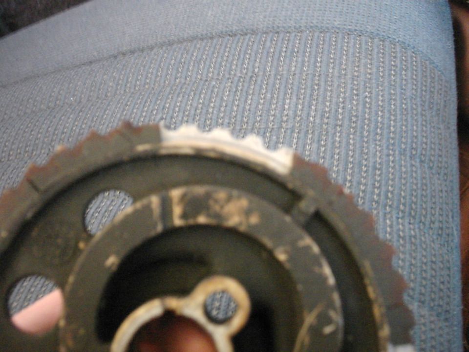 Close-up of a metal timing gear with teeth, showing wear and some rust. A blue fabric surface is visible in the background