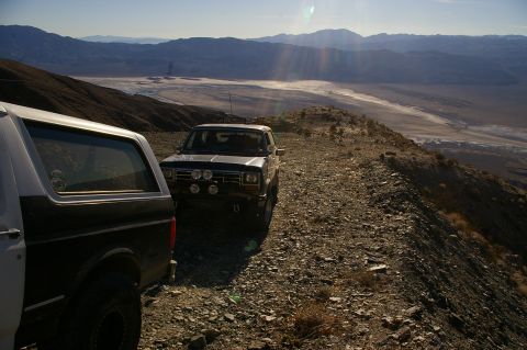 1986 Ford Bronco - Two SUVs are parked on a rocky mountain ridge, with a vast desert valley stretching out below under a clear sky.