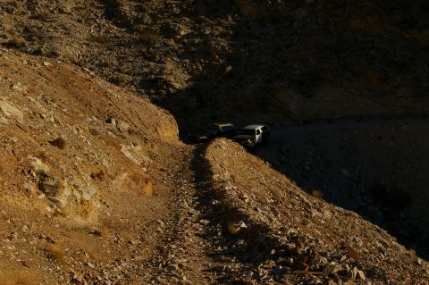 1986 Ford Bronco - Two SUVs navigate a rough dirt road in a desolate, rocky canyon landscape.