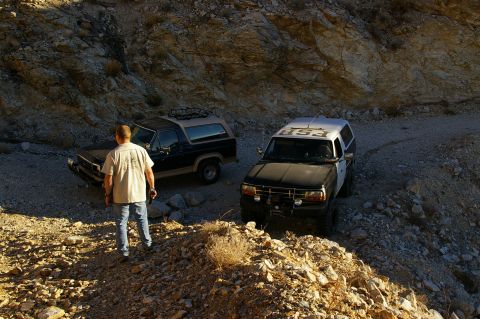 1986 Ford Bronco - Two Ford Broncos are parked on a rocky dirt road in a desert canyon.