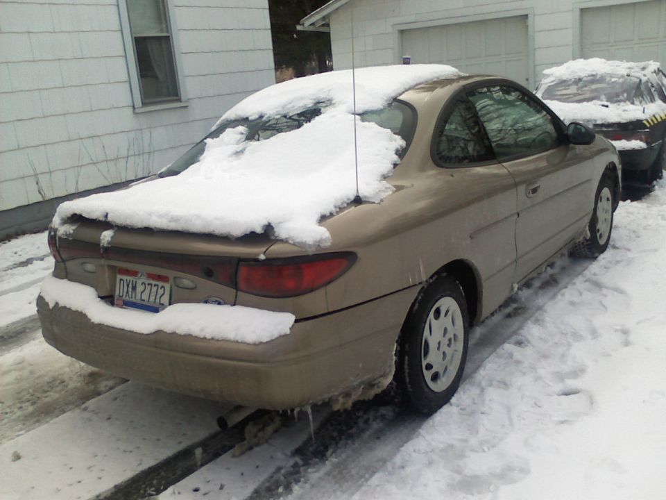 Tan coupe covered in snow, rear view. License plate DXM 2772. Another vehicle is partially visible behind it