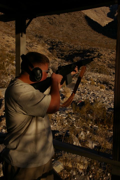 1986 Ford Bronco - A man wearing ear protection and glasses aims a rifle at a desert shooting range.