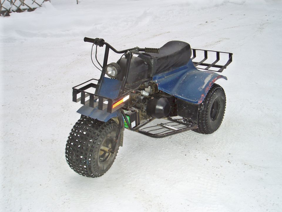 Blue three-wheeled ATV with knobby tires, front basket, and rear rack, parked in snow