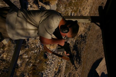 1986 Ford Bronco - Man aims rifle at outdoor shooting range with rocky terrain and sparse vegetation.
