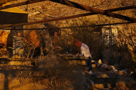 1986 Ford Bronco - People work on something outdoors in a dry, brushy environment under a rustic overhead structure.