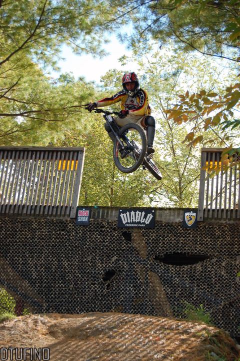 1976 Toyota Land Cruiser - A mountain biker performs a jump over a wooden ramp, with trees and a blue sky visible.