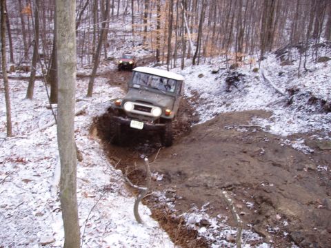 1976 Toyota Land Cruiser - An off-road vehicle navigates a challenging muddy and snowy path through a wooded area.