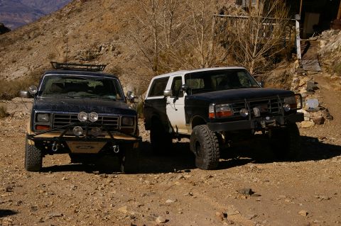 1986 Ford Bronco - Two SUVs are parked side-by-side on a dusty, arid terrain with sparse vegetation and hills in the background.
