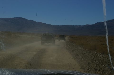 1986 Ford Bronco - Two SUVs drive down a dusty desert road with mountains in the background.